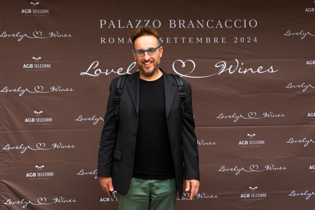 Un uomo con occhiali, blazer nero e zaino sorride davanti a un fondale marrone con le scritte Lovely Wines, PALAZZO BRANCACCIO e ROMA SETTEMBRE 2024.
