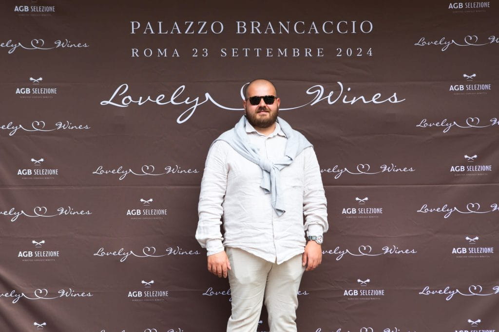 Un uomo con occhiali da sole, camicia bianca e pantaloni chiari si trova davanti a un fondale marrone con testo bianco che recita PALAZZO BRANCACCIO ROMA 23 SETTEMBRE 2024 e Lovely Wines.