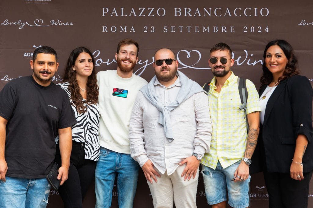 Sei persone sono in piedi, sorridenti, davanti a uno sfondo con la scritta "Palazzo Brancaccio, Roma 23 settembre 2024, Lovely Wines". Sono vestiti in modo casual e posano per una foto di gruppo durante un evento.