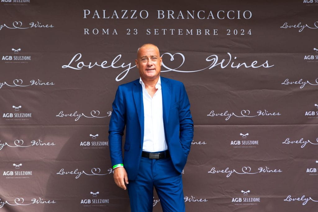 Un uomo in abito blu si trova davanti a un fondale marrone con la scritta Lovely Wines e Palazzo Brancaccio, Roma 23 Settembre 2024 durante un evento.