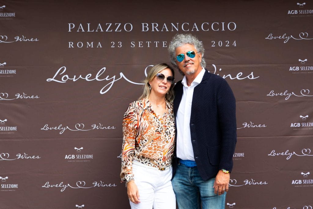 Una coppia sorridente posa davanti a uno striscione a passo di danza per un evento enologico a Palazzo Brancaccio, a Roma, il 23 settembre 2024. L'uomo indossa occhiali da sole; la donna indossa una camicetta a fantasia e pantaloni bianchi.