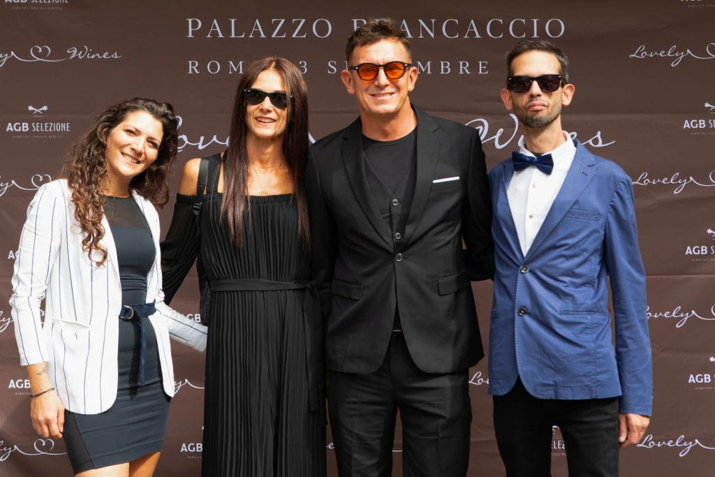 Quattro persone in piedi, una accanto all'altra, sorridono a un evento. Due donne indossano abiti neri (una con un blazer bianco) e due uomini indossano occhiali da sole e abiti scuri, uno nero e uno blu, davanti a uno sfondo di un evento di marca.