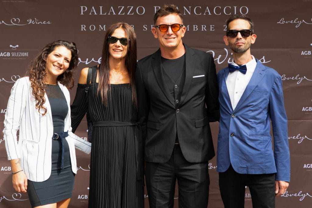 Quattro persone vestite in modo formale stanno insieme e sorridono davanti a uno sfondo con testo in occasione di un evento o di una riunione all'aperto. Il gruppo appare allegro ed elegante, con due uomini in abito e occhiali da sole e due donne in abito.
