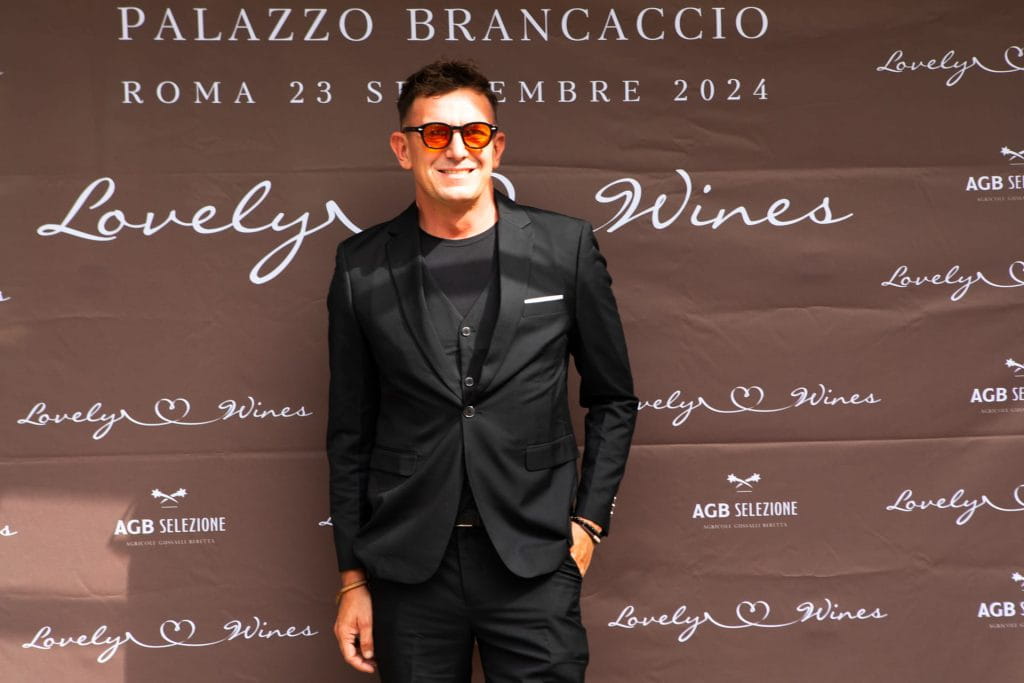 Un uomo in abito nero e occhiali da sole sorride davanti a un fondale marrone con testo bianco che promuove Lovely Wines a Palazzo Brancaccio a Roma, datato 23 settembre 2024.