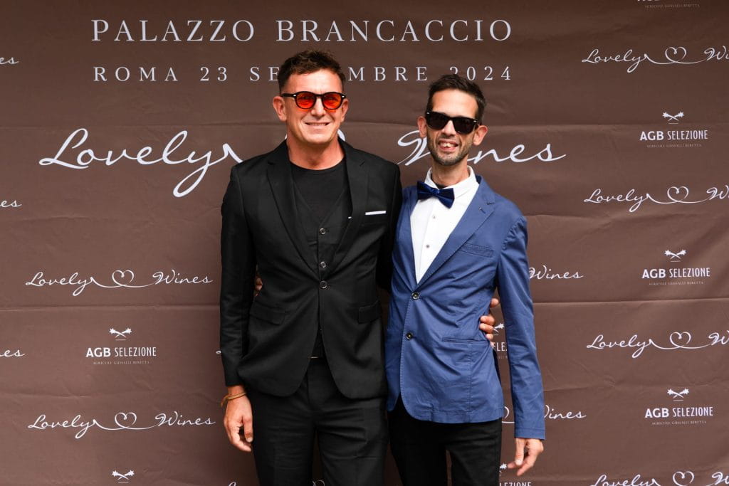 Due uomini vestiti in modo formale posano davanti a un fondale marrone per eventi con testo bianco che recita PALAZZO BRANCACCIO, ROMA 23 SETTEMBRE 2024 e Lovely Wines. Un uomo indossa un abito nero e l'altro un abito blu.