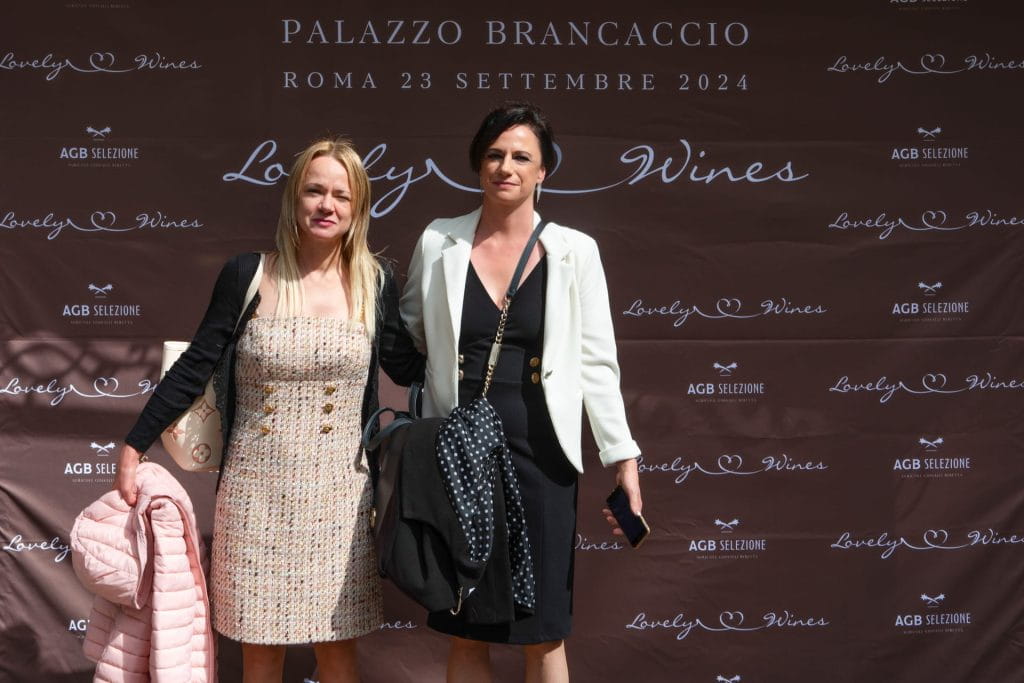 Due donne in piedi e in posa per una foto davanti a un fondale marrone con testo bianco che promuove "Lovely Wines" a Palazzo Brancaccio a Roma, datata 23 settembre 2024. Entrambe tengono una giacca e indossano un abito.