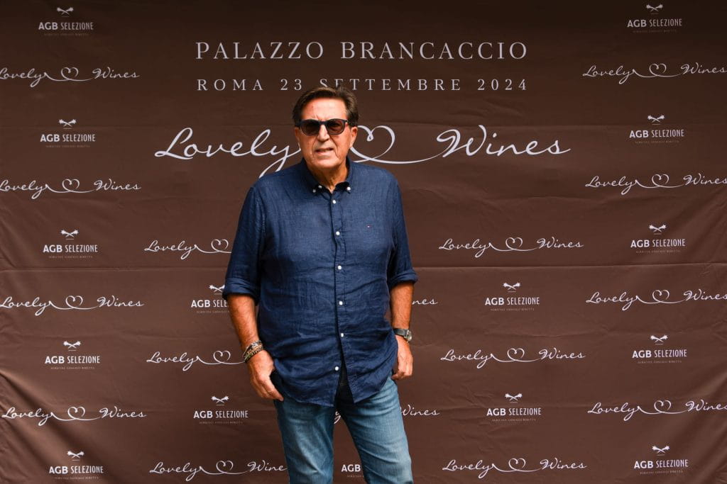 Un uomo con occhiali da sole e camicia blu posa davanti a un fondale marrone con testo bianco che promuove "Lovely Wines", la sede di Palazzo Brancaccio e la data dell'evento, il 23 settembre 2024, a Roma.