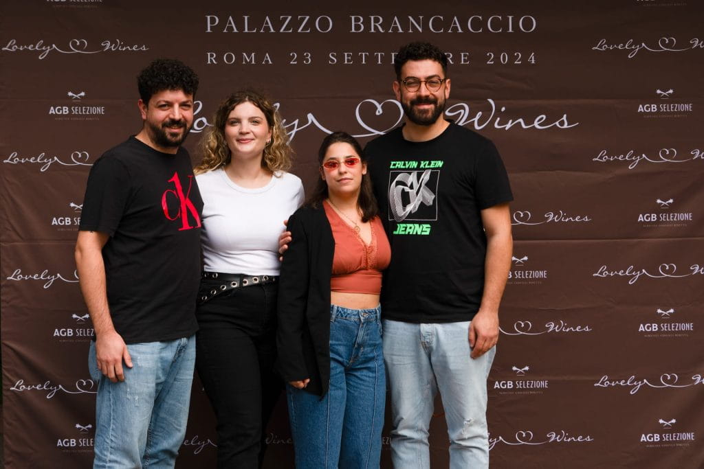 Quattro persone sono in piedi, sorridenti, davanti a un fondale marrone con la scritta Palazzo Brancaccio, Roma 23 Settembre 2024 e Lovely Wines.