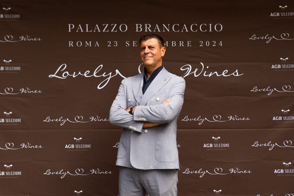 Un uomo in abito grigio chiaro sta in piedi con le braccia incrociate, sorridendo davanti a un fondale marrone con testo bianco che promuove un evento enologico a Palazzo Brancaccio a Roma il 23 settembre 2024.