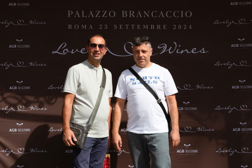 Due uomini posano davanti a uno sfondo marrone con la scritta Palazzo Brancaccio, Roma 23 settembre 2024, Lovely Wines. Entrambi sono vestiti in modo casual e sono in piedi alla luce del sole.