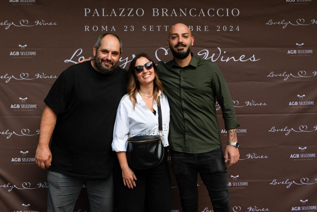 Tre persone in posa e sorridenti davanti a un fondale marrone con un testo che promuove un evento enologico a Palazzo Brancaccio a Roma il 23 settembre 2024. Il gruppo è composto da due uomini e una donna in piedi.
