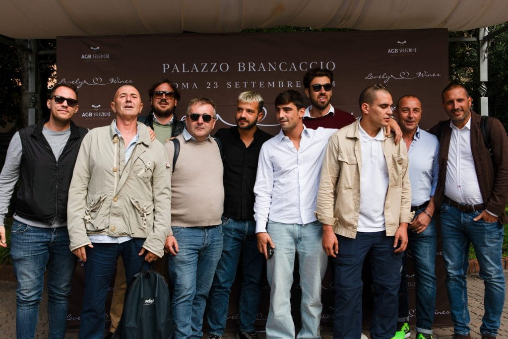 Un gruppo di uomini posa insieme davanti a uno sfondo con la scritta Palazzo Brancaccio Roma 23 Settembre durante un evento all'aperto. Sono vestiti in modo casual e semi-formale e sorridono per la foto.