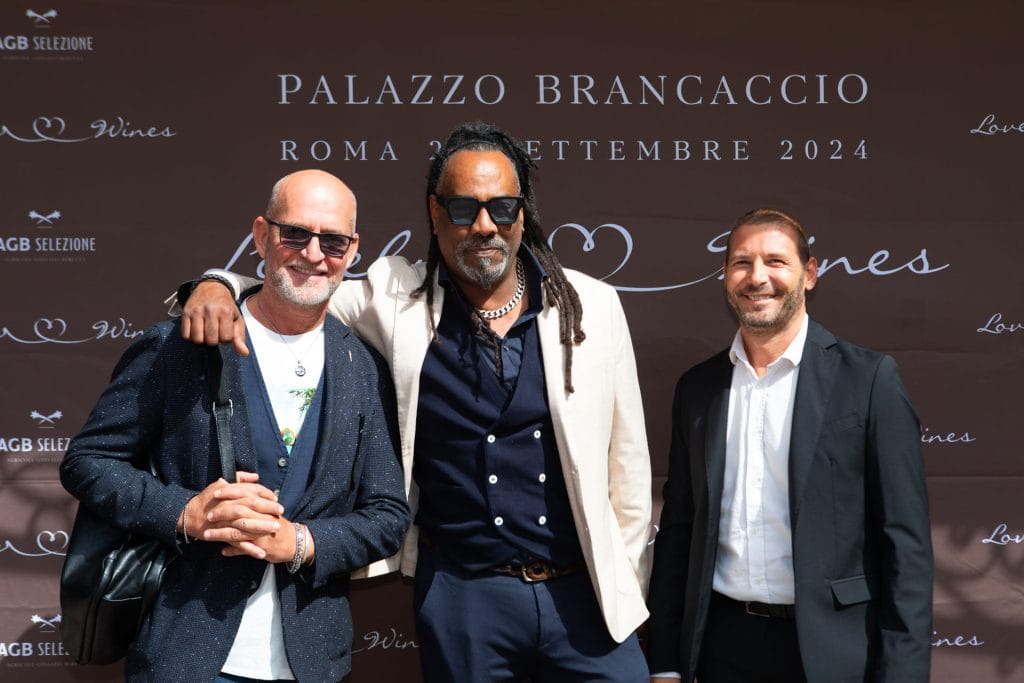 Tre uomini in giacca e cravatta e occhiali da sole sorridono davanti a un fondale marrone con la scritta PALAZZO BRANCACCIO ROMA 24 SETTEMBRE 2024 stampata in alto.