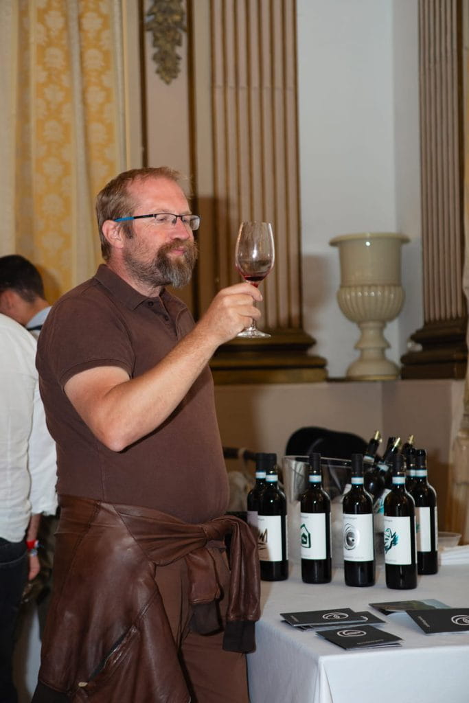 Un uomo con barba e occhiali regge un bicchiere di vino rosso e lo esamina. È in piedi accanto a un tavolo con diverse bottiglie di vino e volantini in una stanza elegante e ben illuminata, con alte colonne e decorazioni ornamentali.