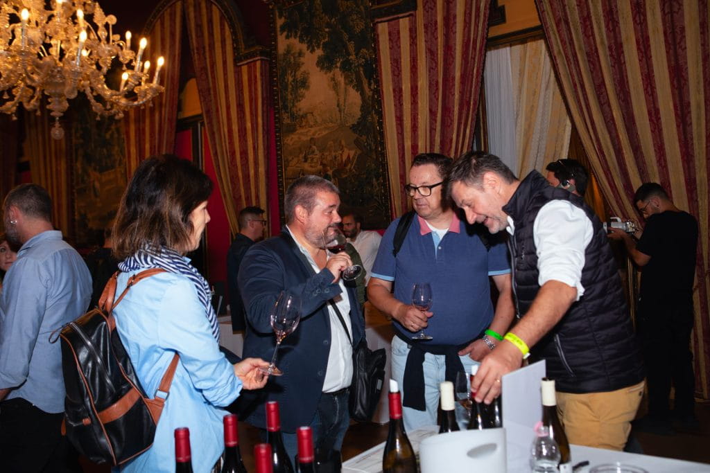 Un gruppo di persone si riunisce intorno a un tavolo con bottiglie di vino durante un evento al chiuso. Un uomo versa il vino, mentre altri tengono i bicchieri e conversano. Sullo sfondo, drappi eleganti e un lampadario decorano la stanza.