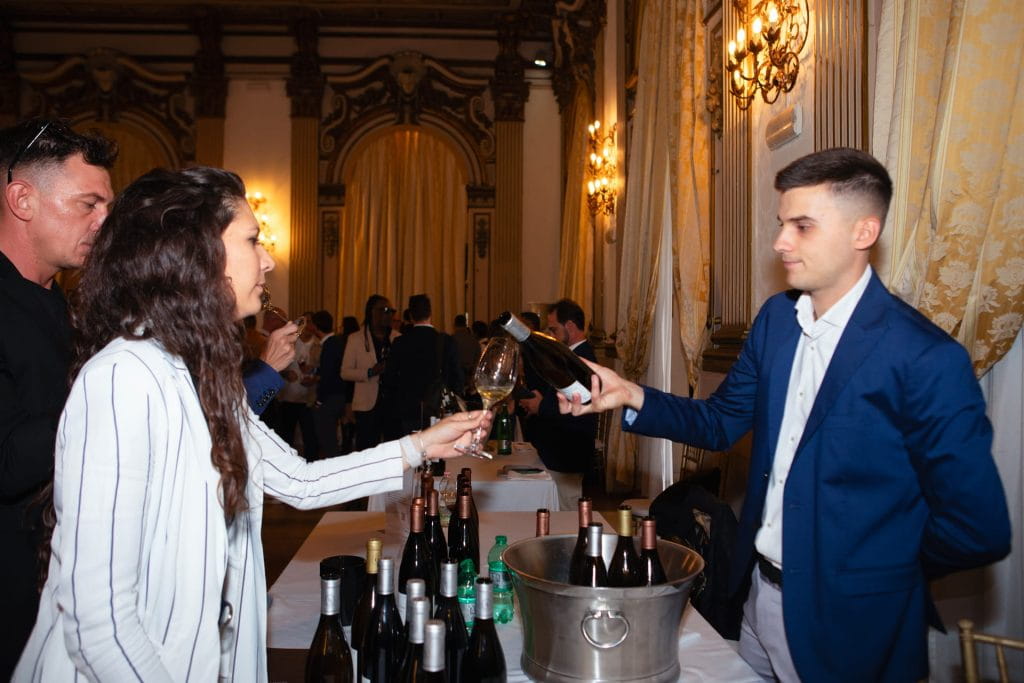 Un uomo in abito blu versa del vino nel bicchiere di una donna durante un elegante evento al chiuso, con bottiglie e un secchiello del ghiaccio sul tavolo; altri ospiti si mescolano sullo sfondo.