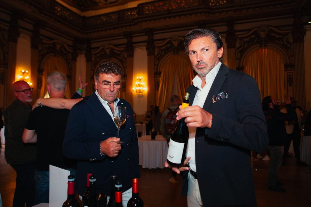 Due uomini in giacca e cravatta assistono a una degustazione di vini in una sala elegante e decorata. Un uomo tiene in mano una bottiglia di vino e guarda la telecamera, mentre l'altro ispeziona un bicchiere di vino. Le bottiglie sono disposte sul tavolo di fronte a loro.