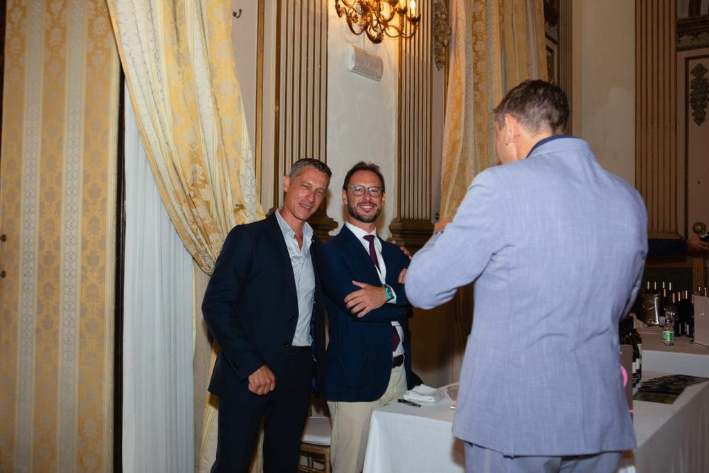 Due uomini in giacca e cravatta posano e sorridono per una foto, mentre un terzo uomo in abito chiaro dà le spalle all'obiettivo in una stanza elegante e decorata con tende e lampadari dorati.