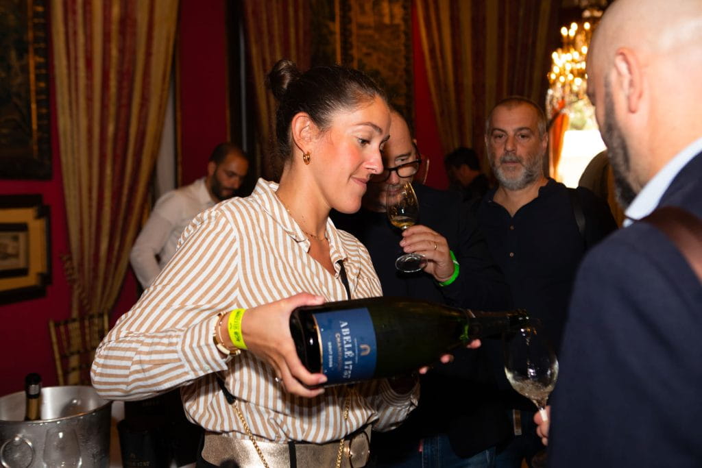 Una donna con una camicia a righe versa del vino da una grande bottiglia in un bicchiere durante un evento al chiuso, con diverse persone intorno a lei che tengono i bicchieri e guardano. Sullo sfondo, tende eleganti e luci calde.