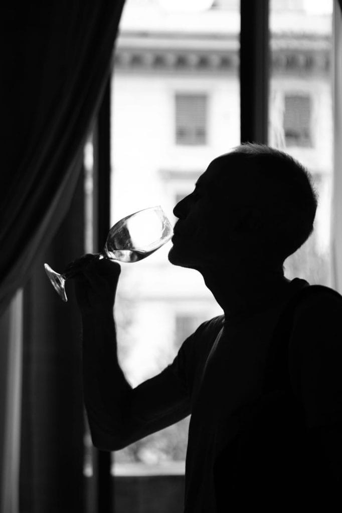 Silhouette di una persona che sorseggia da un bicchiere di vino davanti a un'ampia finestra illuminata dalla luce naturale e con un edificio sfocato visibile all'esterno.