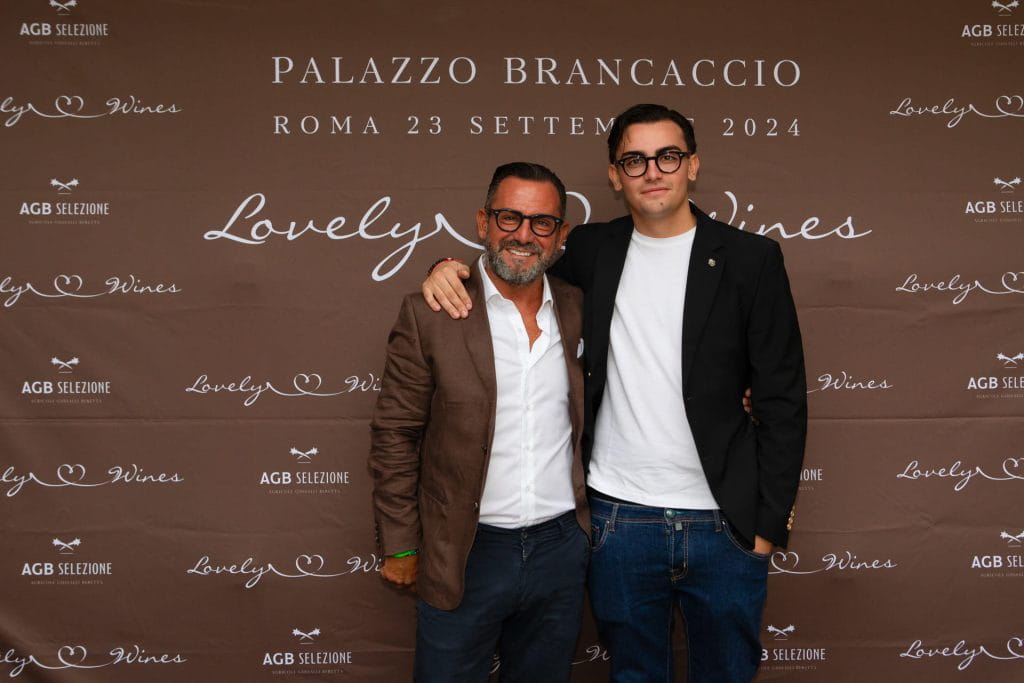 Due uomini sono a braccetto, sorridendo alla telecamera, davanti a un fondale marrone con la scritta bianca PALAZZO BRANCACCIO ROMA 23 SETTEMBRE 2024 e i loghi di Lovely Wines e AGB SELEZIONE.