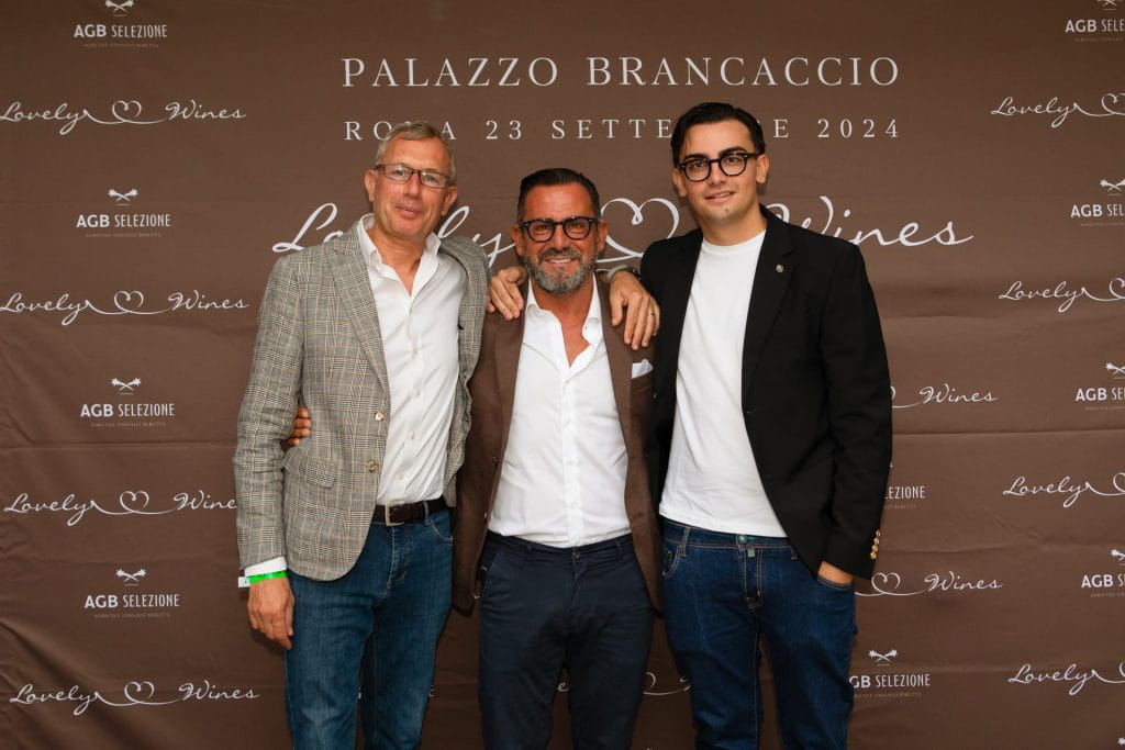 Tre uomini posano insieme, sorridenti, davanti a un fondale marrone con testo bianco che recita Palazzo Brancaccio, Roma 23 Settembre 2024, e Lovely Wines. Sono vestiti in modo elegante e casual.