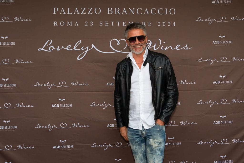 Un uomo con occhiali da sole, giacca di pelle nera e jeans è in piedi sorridente davanti a un fondale marrone con le scritte Palazzo Brancaccio, Roma 23 Settembre 2024 e Lovely Wines.