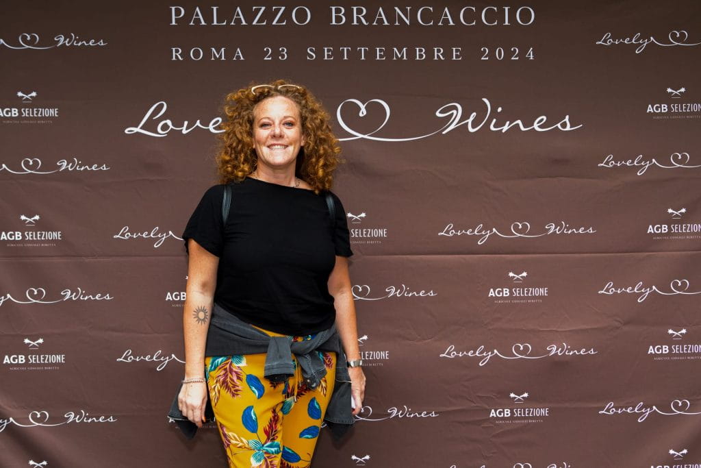 Una donna con i capelli ricci, che indossa un top nero e pantaloni colorati, si trova sorridente davanti a un fondale marrone con la scritta Love Wines, Palazzo Brancaccio e la data dell'evento Roma 23 Settembre 2024.