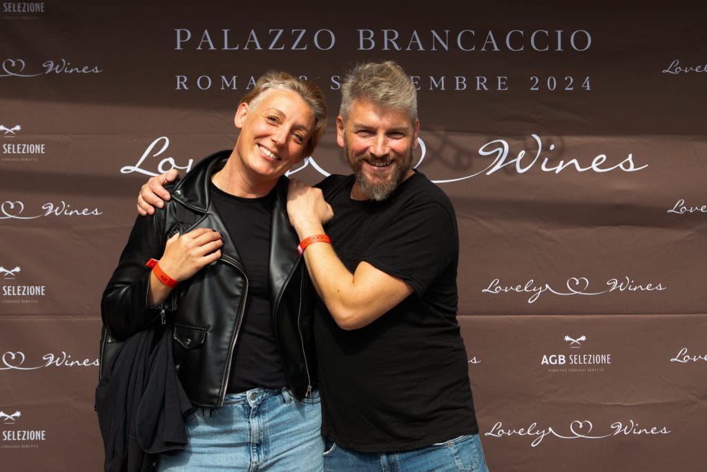 Due persone sorridono e posano insieme davanti a un fondale marrone con scritte bianche PALAZZO BRANCACCIO e Lovely Wines. Entrambi indossano abiti casual e braccialetti rossi. L'evento sembra svolgersi a Roma, nel settembre 2024.