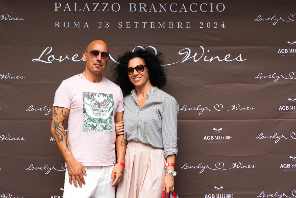 Un uomo in maglietta chiara e pantaloncini sta accanto a una donna in camicetta grigia e gonna rosa, entrambi sorridenti davanti a un fondale marrone con i dettagli dell'evento "Lovely Wines" e "Palazzo Brancaccio".