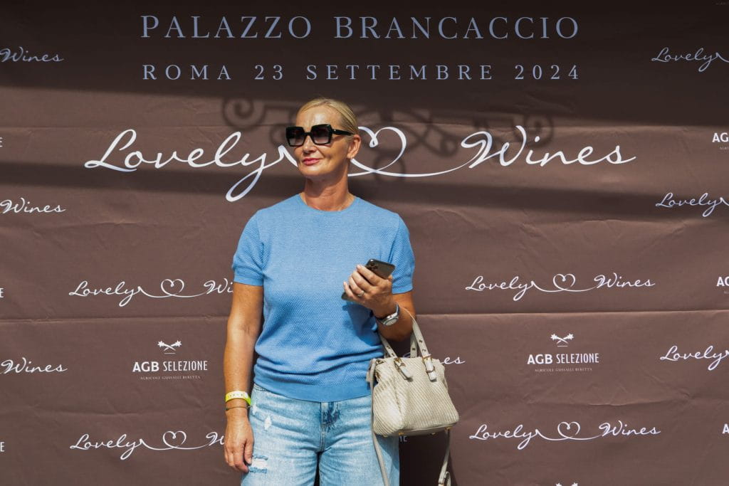 Una donna con occhiali da sole e camicia blu tiene in mano un telefono e una borsa beige davanti a un fondale con la scritta "Lovely Wines" che annuncia un evento a Palazzo Brancaccio il 23 settembre 2024, a Roma.