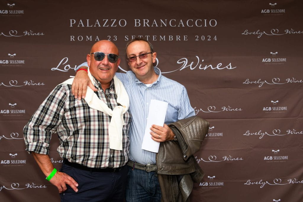 Due uomini sorridono e posano insieme davanti a un fondale con la scritta "Palazzo Brancaccio, Roma 23 settembre 2024" e vari loghi legati al vino. Uno dei due uomini indossa occhiali da sole; entrambi appaiono rilassati e felici.