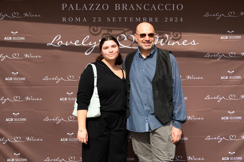 Una giovane donna e un uomo anziano stanno insieme sorridendo davanti a un fondale marrone con testo bianco per Lovely Wines e Palazzo Brancaccio, Roma 23 settembre 2024.