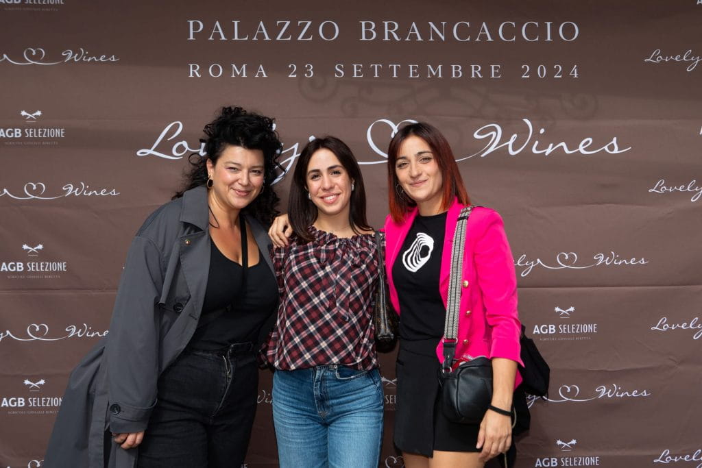 Tre donne stanno insieme sorridendo davanti a un fondale marrone per eventi con il testo "PALAZZO BRANCACCIO ROMA 23 SETTEMBRE 2024" e "Lovely Wines". Sono vestite in modo casual e sembrano felici.
