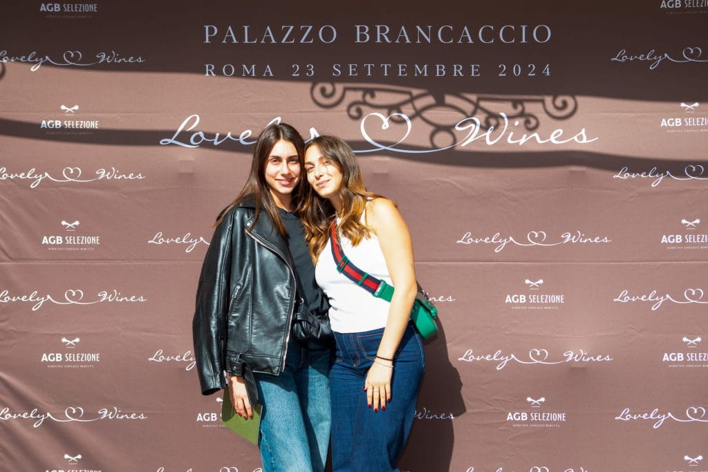 Due donne posano e sorridono insieme davanti a uno sfondo brandizzato per Lovely Wines a Palazzo Brancaccio, Roma, il 23 settembre 2024. Una indossa una giacca nera, l'altra un top bianco e una borsa verde.