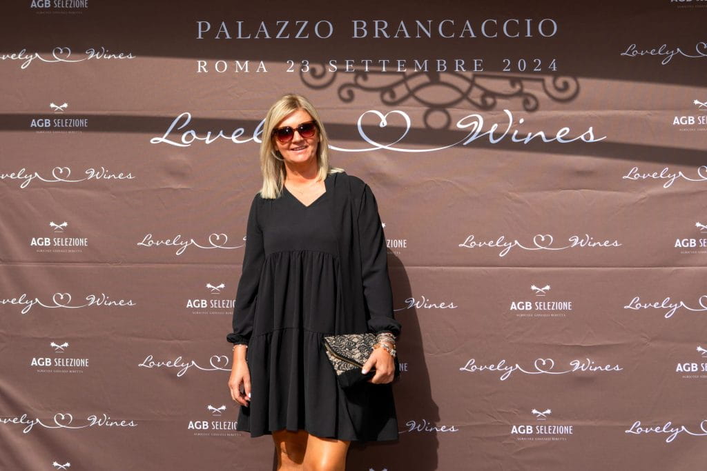 Una donna in abito nero e occhiali da sole sorride davanti a un fondale con Lovely Wines, dettagli dell'evento e testi decorativi a Palazzo Brancaccio, Roma, 23 settembre 2024.