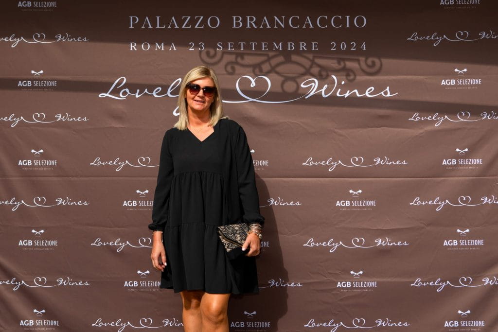 Una donna in abito nero e occhiali da sole sorride davanti a un fondale marrone che promuove i vini Lovely a Palazzo Brancaccio, Roma, 23 settembre 2024.