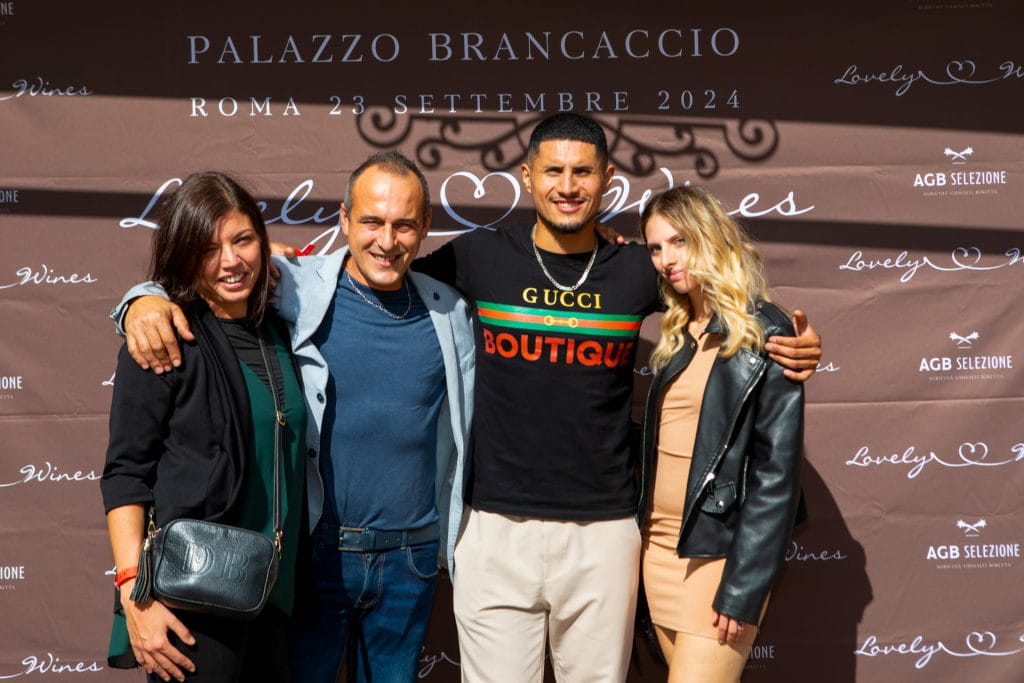 Quattro persone, due uomini e due donne, sorridono abbracciati davanti a un fondale marrone con stampato Palazzo Brancaccio e i dettagli dell'evento. Il gruppo appare felice e vestito in modo elegante e casual.