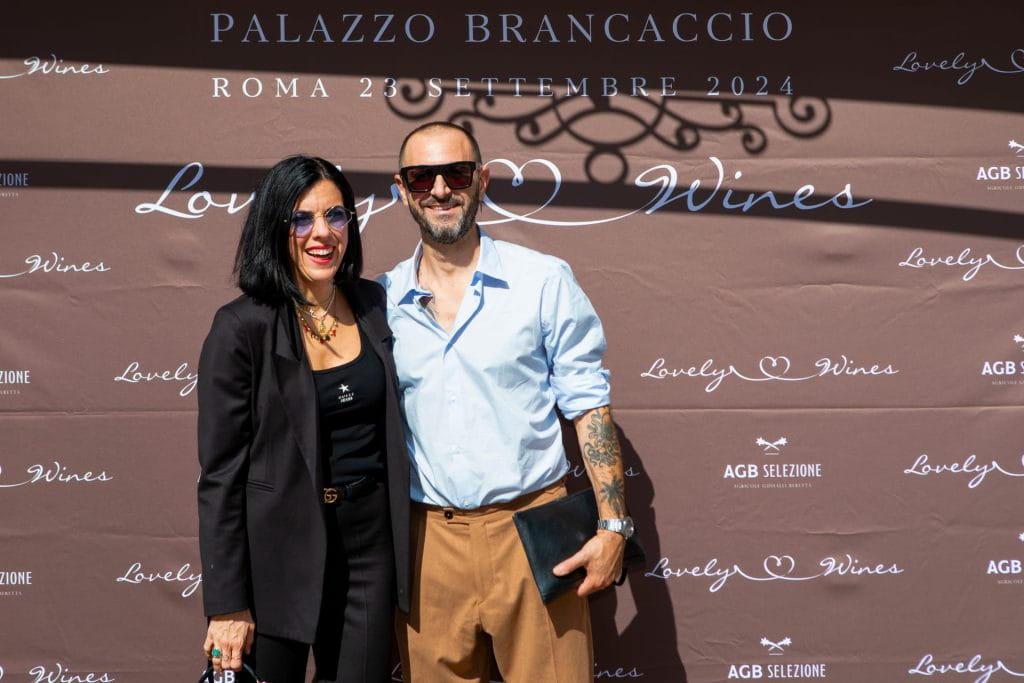 Una donna e un uomo posano sorridenti davanti a uno sfondo marrone con la scritta "Palazzo Brancaccio, Roma 23 settembre 2024, Lovely Wines". Entrambi indossano occhiali da sole con stile.