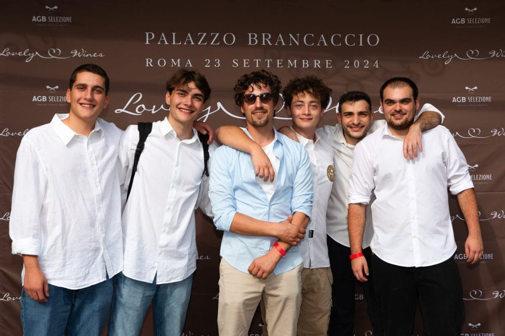 Sei giovani uomini, tutti vestiti con camicie bianche e sorridenti, stanno insieme davanti a un fondale marrone che recita "Palazzo Brancaccio, Roma 23 Settembre 2024, Lovely Wines".