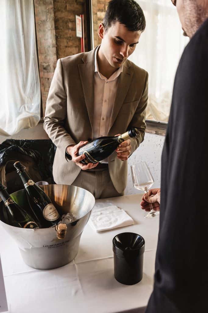 Un uomo in abito marrone versa del vino da una bottiglia in un bicchiere tenuto in mano da un'altra persona a un tavolo con un secchiello del ghiaccio in metallo contenente bottiglie di vino. Sul tavolo si trovano un tovagliolo bianco e un contenitore nero.