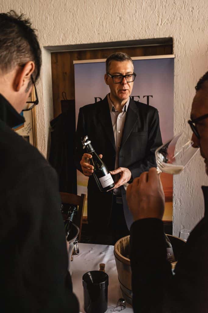 Un uomo con occhiali e giacca scura è in piedi a un tavolo, tiene in mano una bottiglia di vino e parla con due persone, una delle quali sta assaggiando il vino da un bicchiere. Sul tavolo ci sono bottiglie e un secchio.