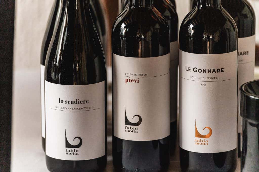 Tre bottiglie di vino rosso con etichette bianche minimaliste su un tavolo. Le etichette recitano lo scudiere, pievi e Le Gonnare, con il marchio di fabio motta e grafiche d'accento nere o arancioni.