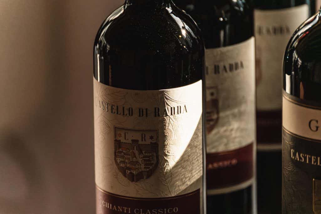 Primo piano di tre bottiglie di vino con eleganti etichette, di cui la più importante è etichettata "Castello di Radda Chianti Classico", con uno stemma dettagliato e una goffratura decorativa, illuminata da una luce calda.