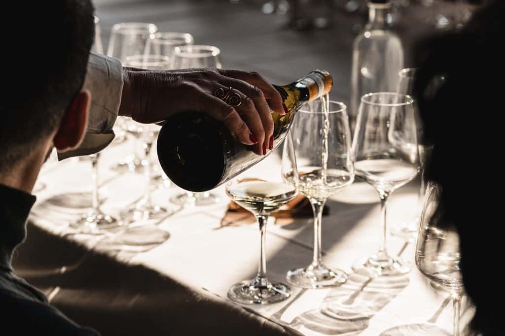 Una persona versa vino bianco da una bottiglia in un bicchiere da vino su un tavolo con diversi bicchieri vuoti e parzialmente riempiti, in una calda luce naturale.