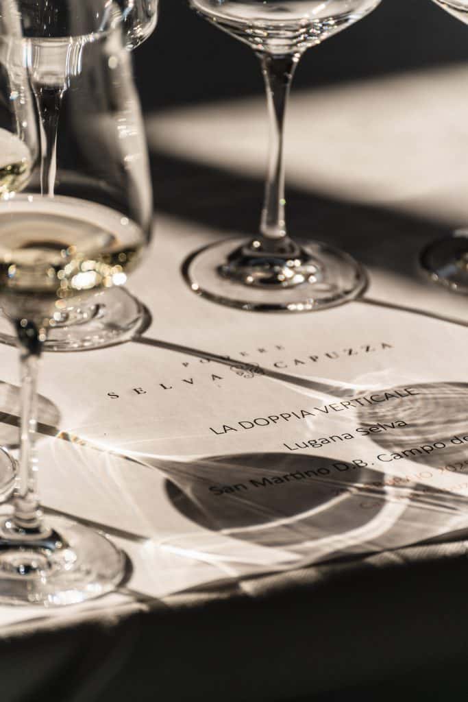 Primo piano di un allestimento per la degustazione di vini, con bicchieri di vino bianco che proiettano ombre su un foglio stampato per l'evento con l'etichetta La Doppia Verticale e Selva Capuzza.