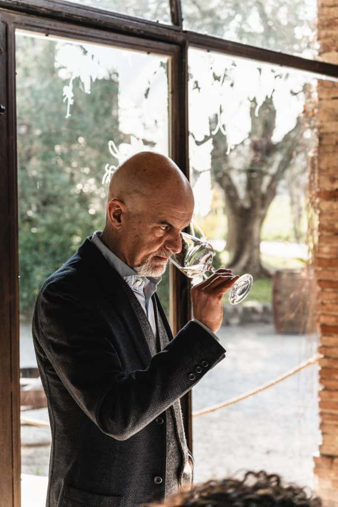 Un uomo in giacca e cravatta è in piedi davanti a una finestra e porta un bicchiere di vino al naso mentre annusa il vino. La luce del sole filtra attraverso la porta a vetri alle sue spalle e gli alberi sono visibili all'esterno.