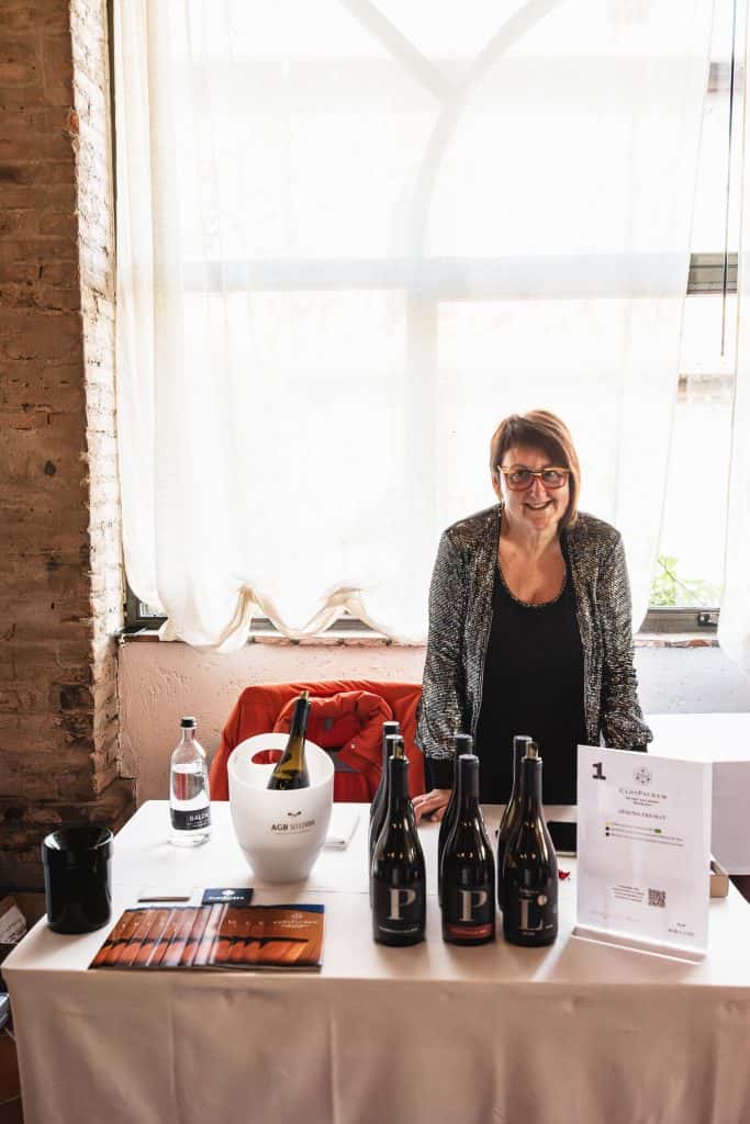 Una donna con gli occhiali è in piedi dietro un tavolo che espone bottiglie di vino, un refrigeratore e opuscoli. È al chiuso, davanti a una grande finestra con tende trasparenti e pareti di mattoni a vista.