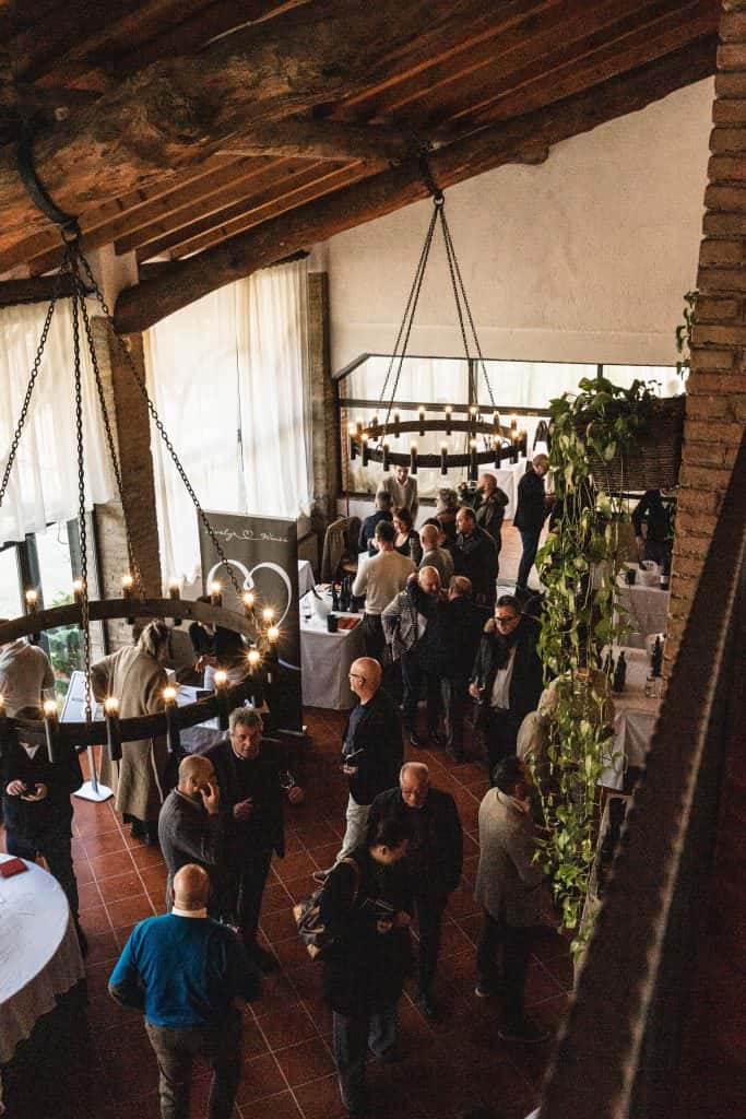 Un gruppo di persone socializza e si mescola in un locale rustico illuminato con travi in legno, pareti in mattoni, grandi finestre e luci decorative appese. Tavoli con tovaglie bianche sono sparsi in tutto lo spazio.
