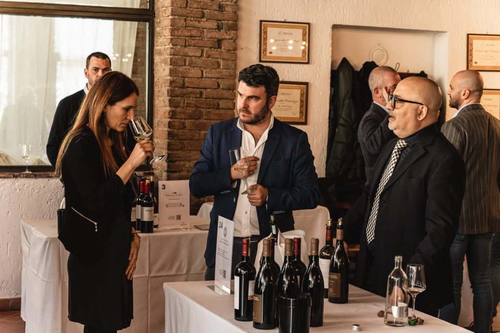 Una donna annusa un bicchiere di vino mentre due uomini osservano un evento di degustazione. Sul tavolo di fronte a loro sono esposte diverse bottiglie di vino. Sullo sfondo sono visibili altre persone.